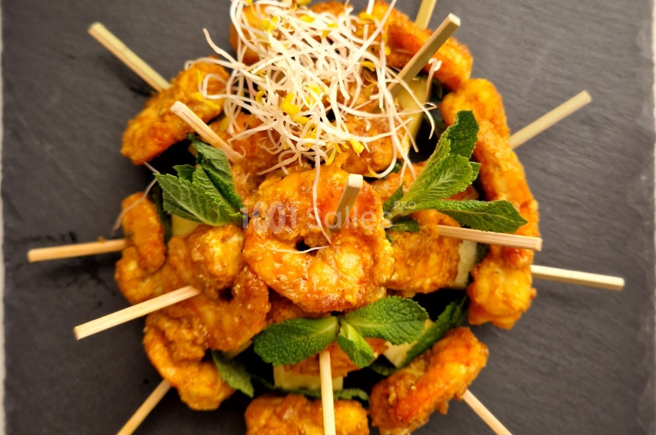 Brochettes de crevettes dorées garnies de feuilles de menthe et de pousses germées, présentées sur une ardoise.