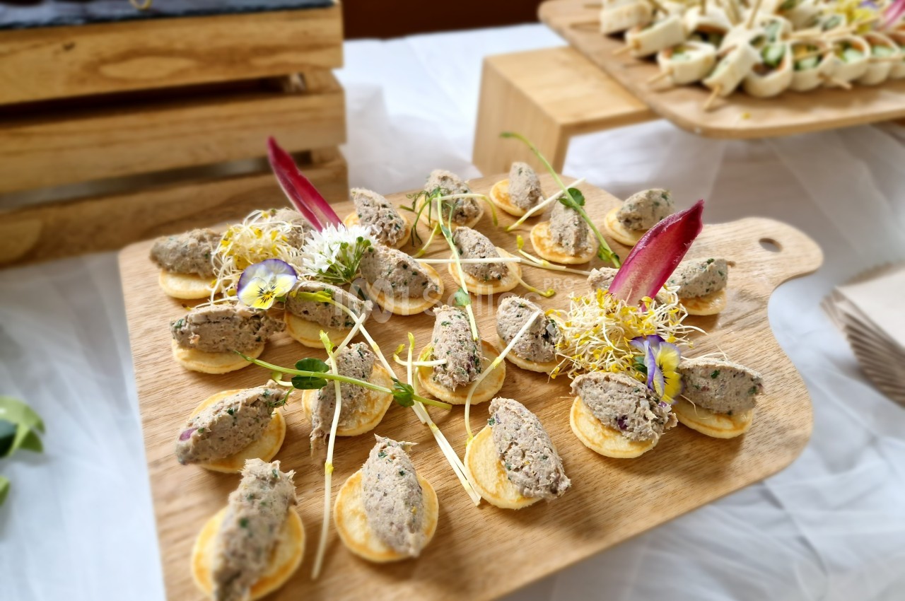 Plateau de mini-blinis garnis de rillettes et décorés de pousses et fleurs comestibles, présenté sur une planche en bois.