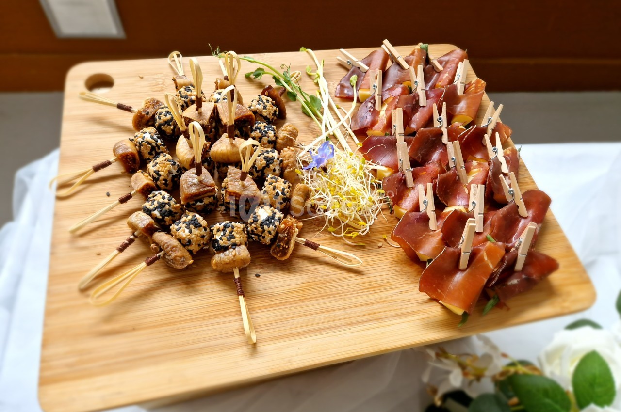 Plateau en bois avec brochettes apéritives de figues et sésame à gauche, jambon cru et fromage à droite.