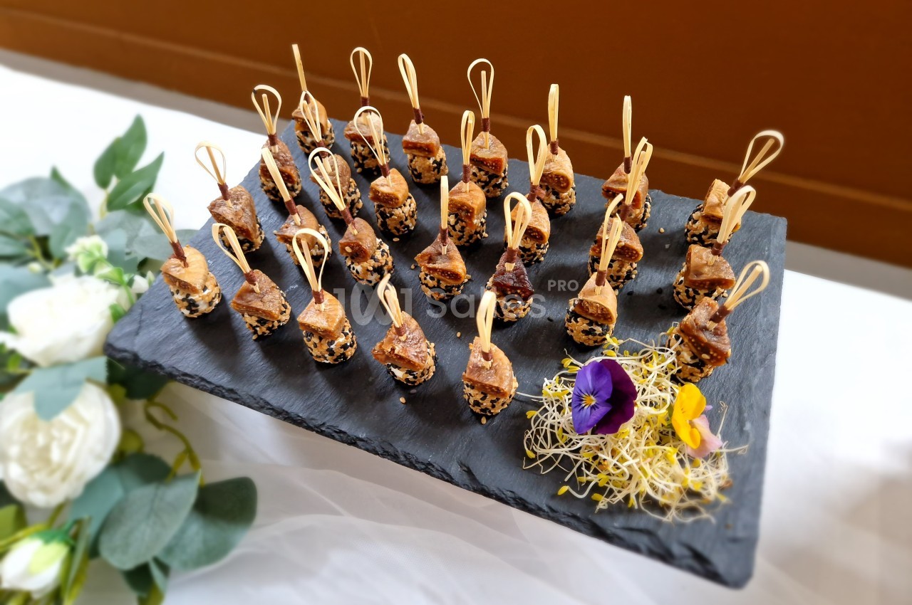 Plateau d'amuse-bouches sur brochettes en bois, disposé sur une ardoise avec décoration florale et végétale.