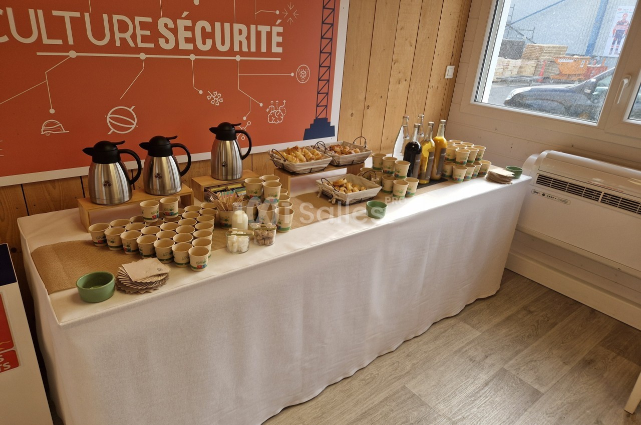 Table dressée avec boissons chaudes, jus, viennoiseries et gobelets dans une salle lumineuse au décor professionnel.