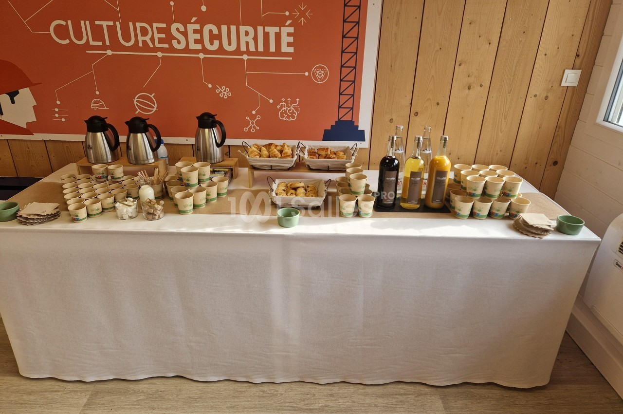 Table dressée avec boissons chaudes, jus, viennoiseries et gobelets, dans une salle avec un panneau ’Culture Sécurité’.