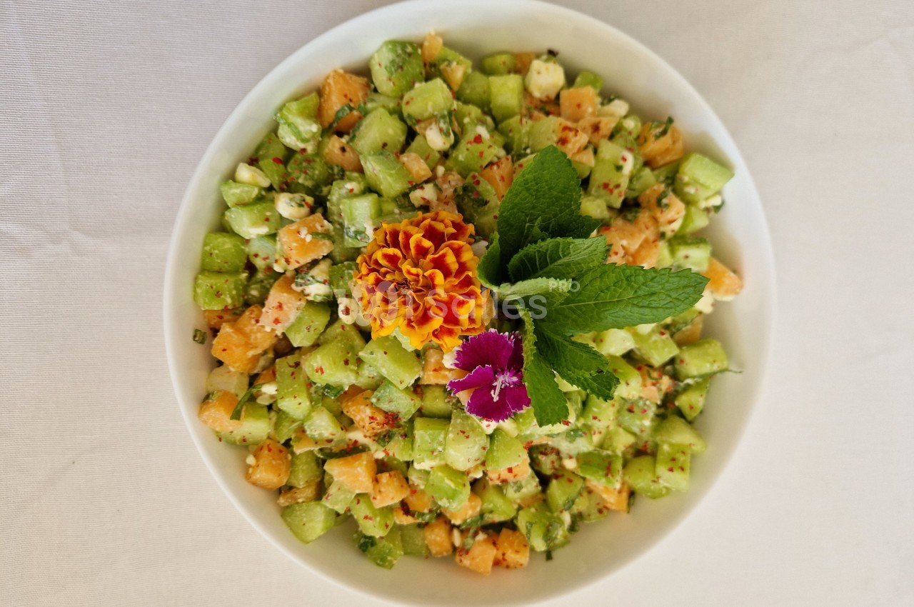 Salade colorée de concombres et melons en dés, garnie de fleurs comestibles et feuilles de menthe sur une nappe blanche.