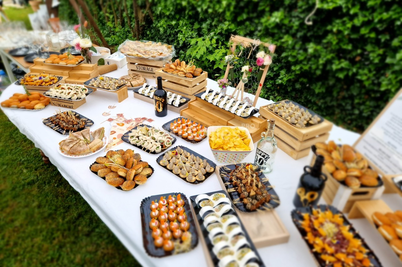 Table garnie de divers amuse-bouches et plats apéritifs disposés sur des plateaux dans un jardin verdoyant.