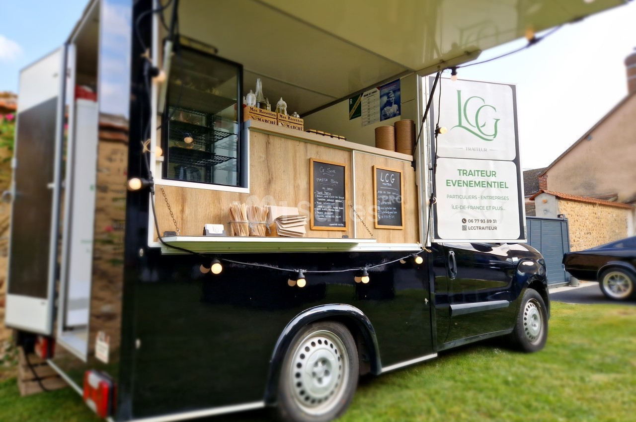 Camion noir aménagé en food truck, avec menu affiché et guirlande lumineuse, stationné sur une pelouse.