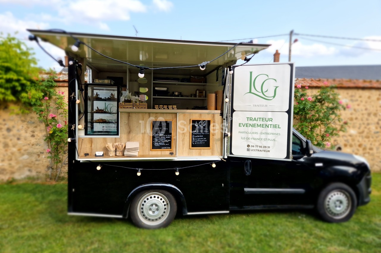 Camion noir aménagé en food truck, avec guirlandes lumineuses et étagères exposant des produits, stationné sur une pelouse.