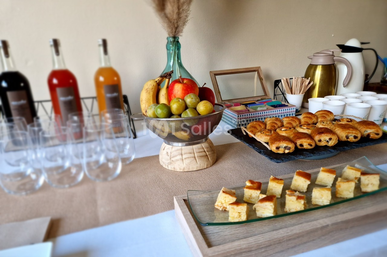 Table dressée avec des boissons, fruits, viennoiseries, cubes de gâteau et tasses pour un buffet ou une collation.