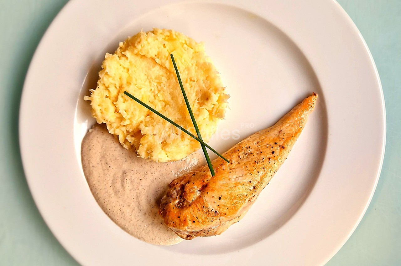 Assiette avec filet de poulet grillé, purée de pommes de terre, sauce crémeuse et brins de ciboulette en décoration.