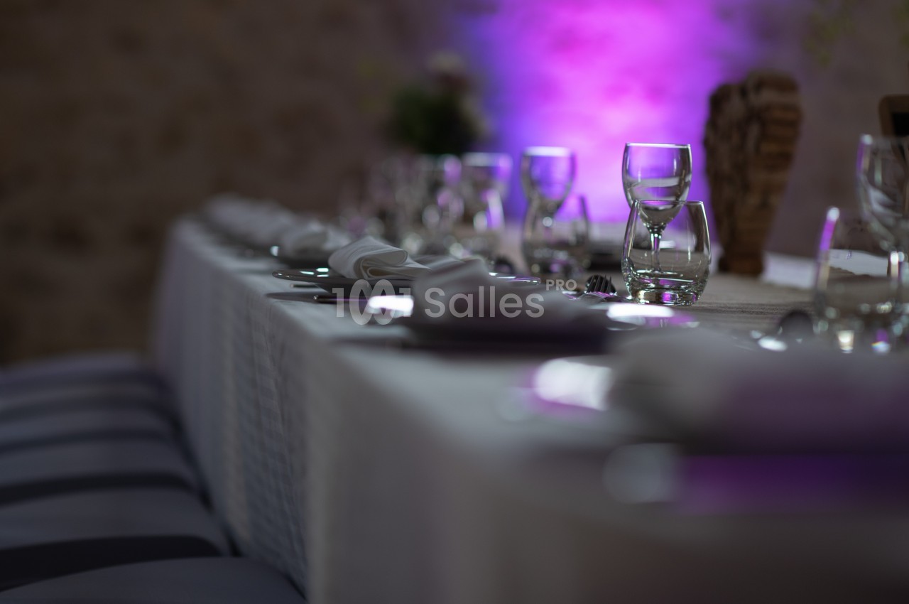 Table dressée avec nappes blanches, verres et serviettes, éclairée par une lumière violette en arrière-plan.