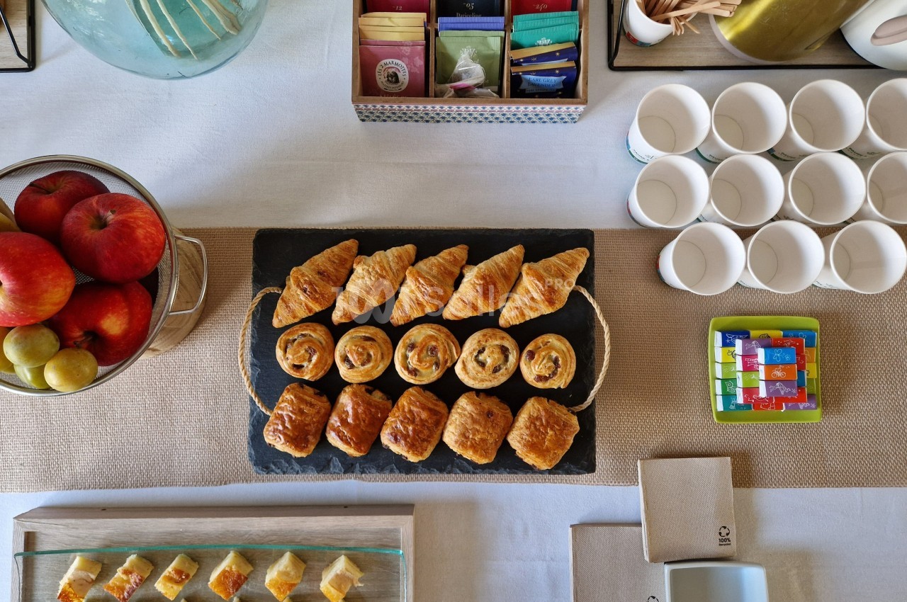 Plateau de viennoiseries variées sur une table dressée avec fruits, tasses et accessoires pour le petit-déjeuner.