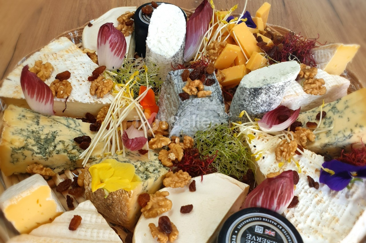 Plateau de fromages variés avec noix, raisins secs, endives, fleurs comestibles et germes, présenté sur un fond en bois.