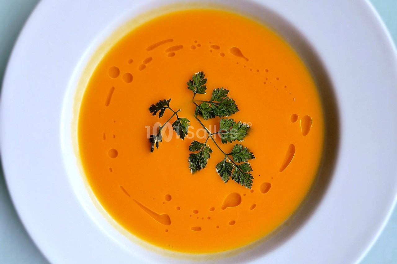 Bol de soupe orange crémeuse, décoré de feuilles de persil et de gouttes d'huile sur une assiette blanche.