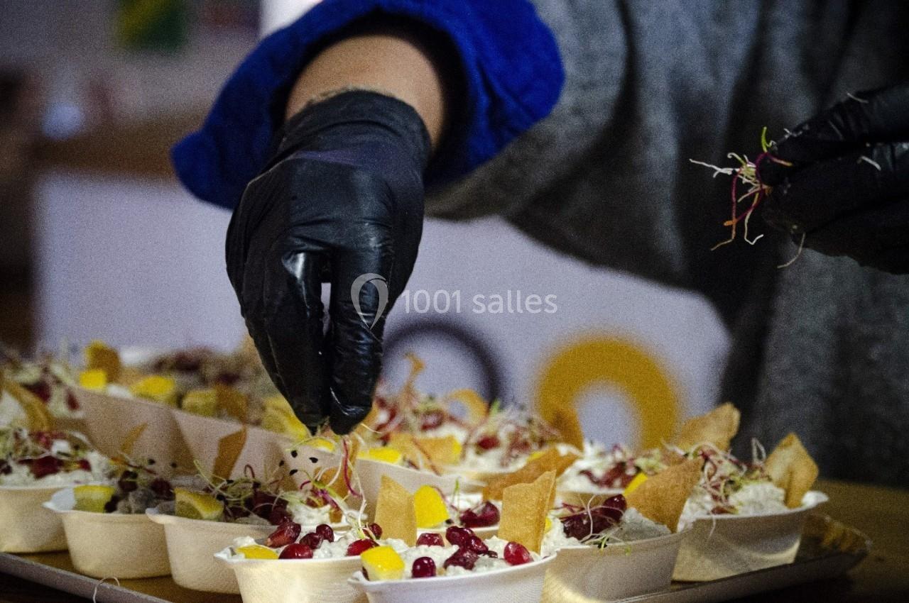 Une personne garnit des plats individuels avec des pousses et des fruits, portant des gants noirs pour l'hygiène.