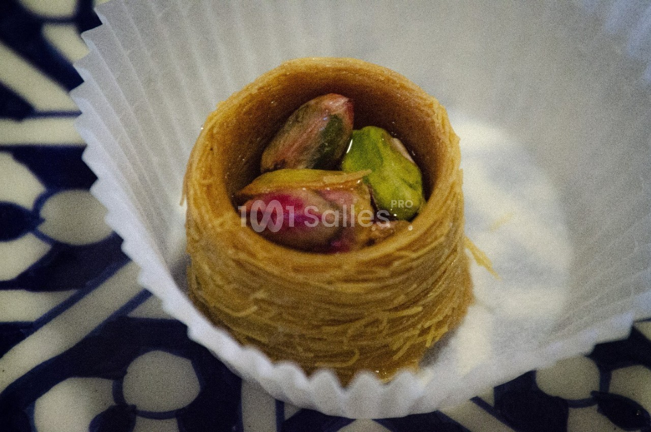 Pâtisserie orientale en nid de kadaïf garnie de pistaches, présentée dans une caissette en papier blanc.