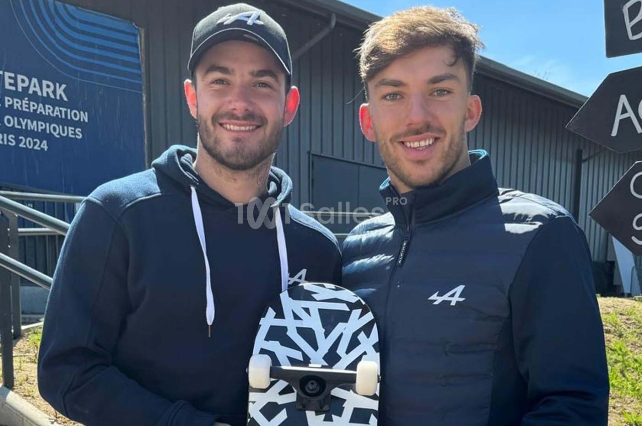 Deux hommes souriants posent à l'extérieur, l'un tenant un skateboard noir et blanc devant un bâtiment moderne.