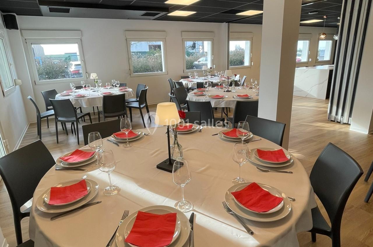 Salle de restaurant lumineuse avec tables rondes dressées, nappes blanches, serviettes rouges et chaises noires.