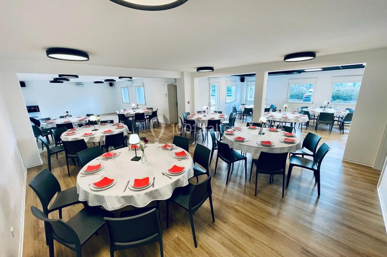 Salle de réception lumineuse avec tables rondes dressées, nappes blanches, serviettes rouges et chaises noires.