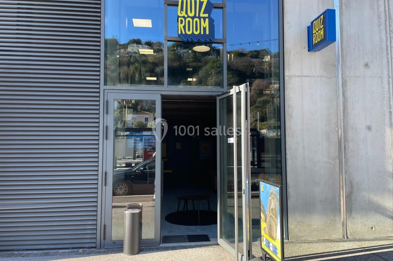 Entrée d'un bâtiment avec une porte vitrée ouverte, une enseigne ’Quiz Room’ et un panneau publicitaire devant.