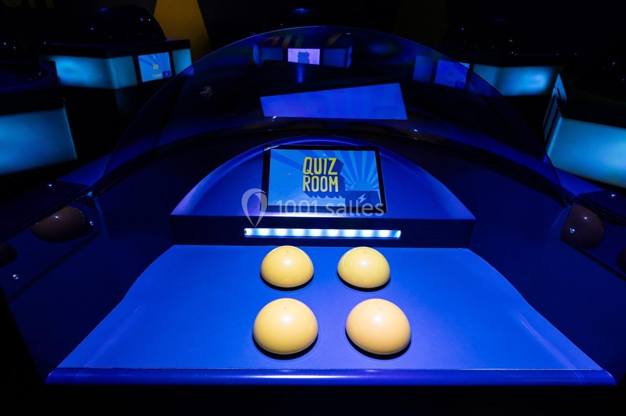 Poste de jeu interactif avec quatre boutons jaunes et un écran affichant ’Quiz Room’ dans un éclairage bleu.