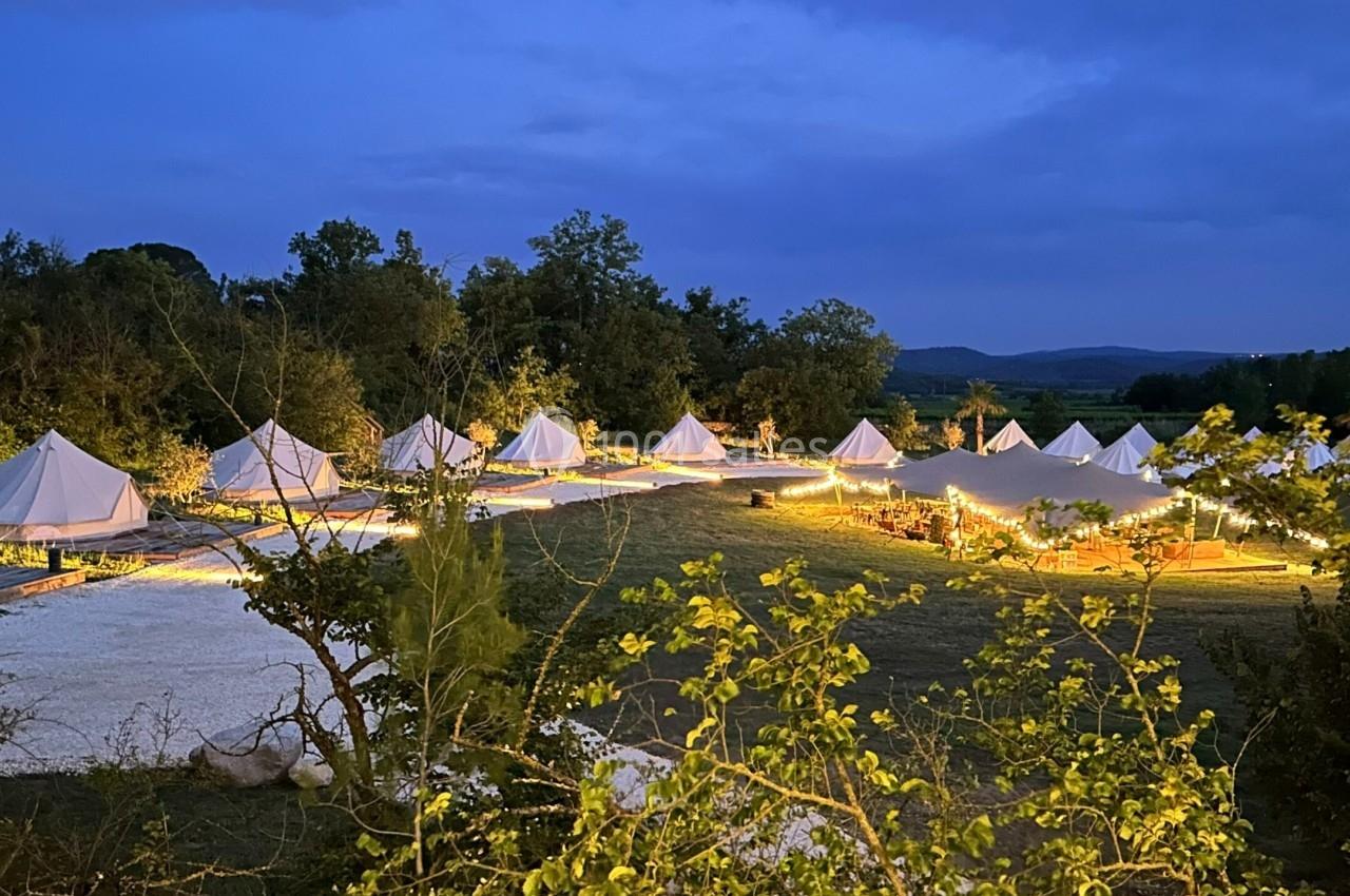 Alignement de tentes blanches éclairées dans un cadre naturel au crépuscule, entouré de végétation et de collines.