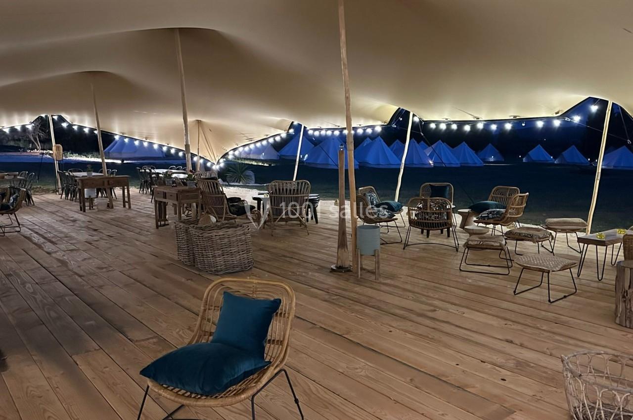 Espace couvert en bois avec des chaises et des tables, éclairé par des guirlandes lumineuses, avec des tentes en arrière…