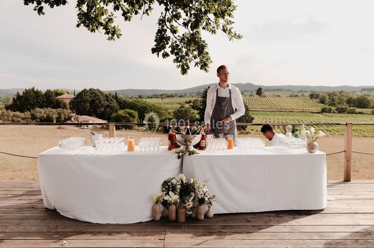 Un serveur derrière une grande table dressée avec des boissons et décorée de fleurs, en extérieur avec vue sur des vignes.