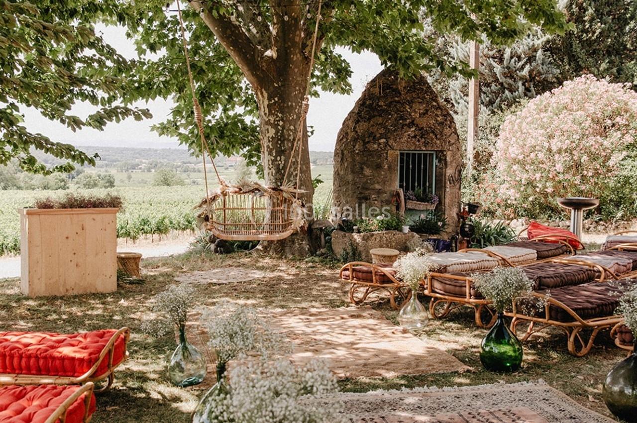 Espace extérieur aménagé avec des fauteuils en rotin, tapis et décorations florales sous un arbre près d'une petite bâtisse.
