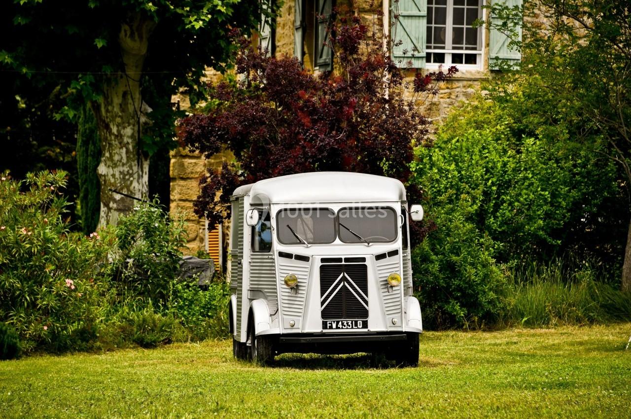 Fourgon Citroën vintage gris stationné sur une pelouse devant une maison en pierre entourée de végétation.