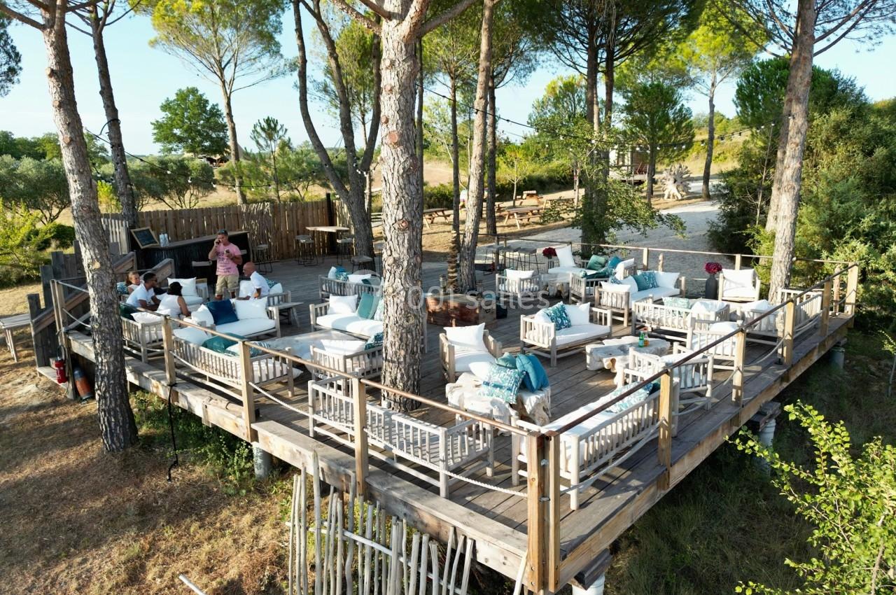 Terrasse en bois surélevée aménagée avec des fauteuils et tables, entourée d'arbres dans un cadre naturel.