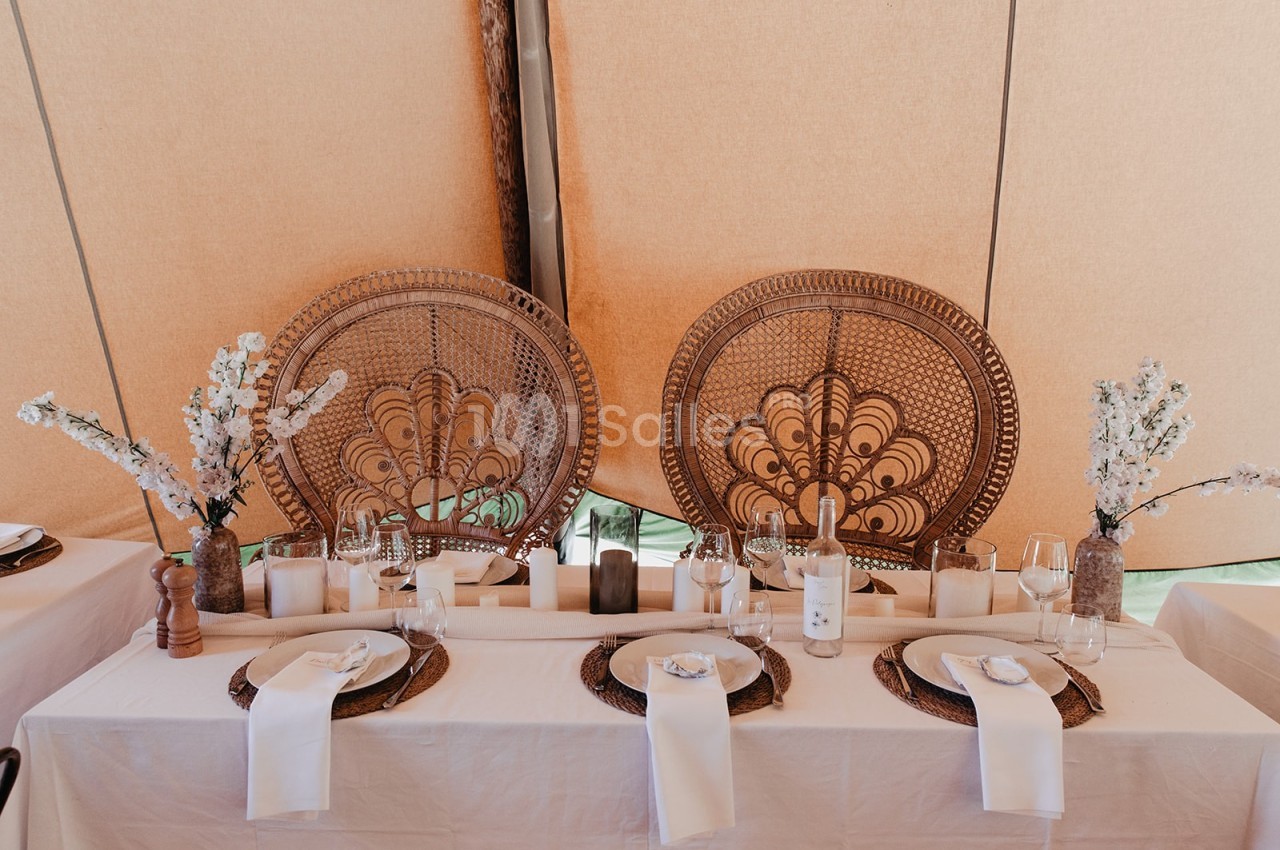 Table décorée avec des assiettes, serviettes blanches, bougies et fleurs, devant des dossiers de chaises en rotin.