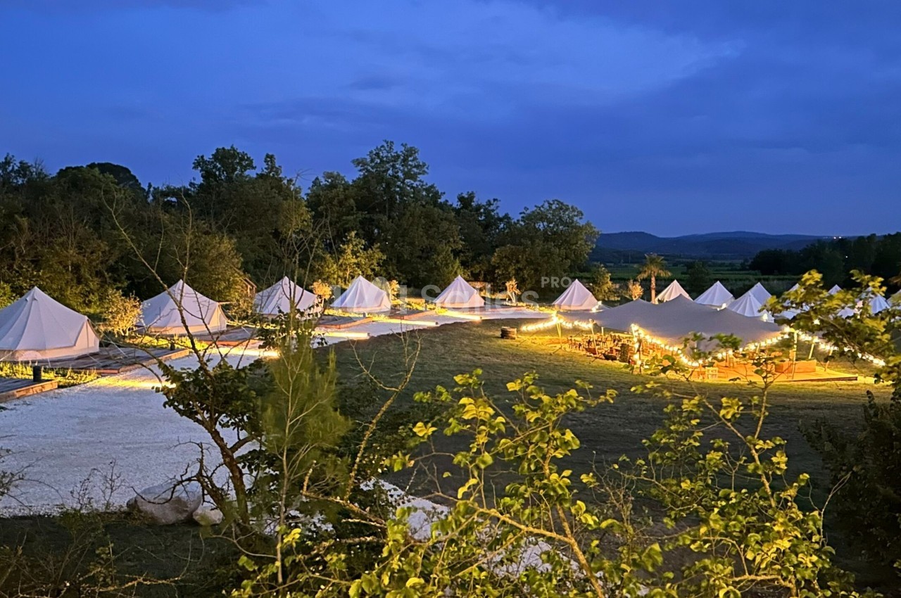 Alignement de tentes blanches éclairées dans un cadre naturel au crépuscule, entouré de végétation et de collines.