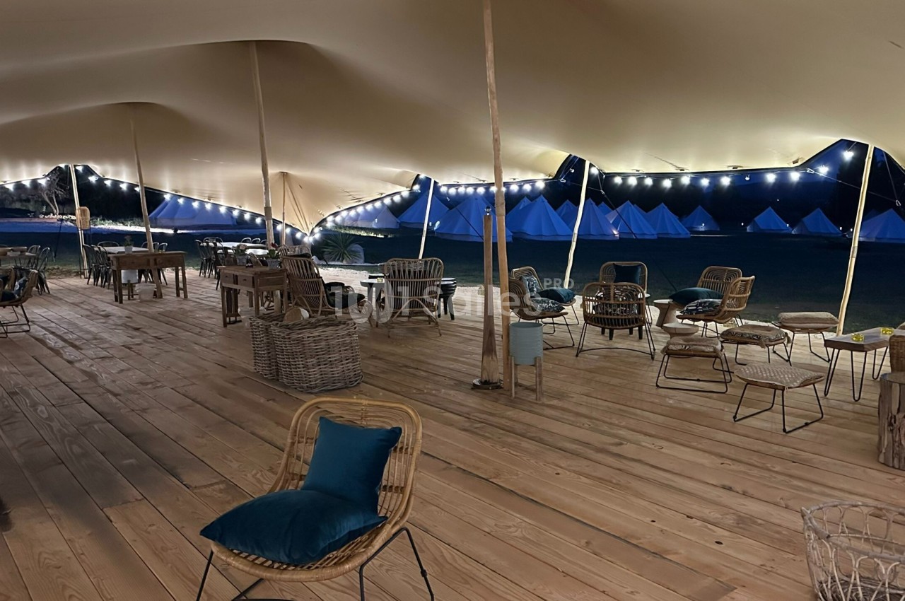 Espace couvert en bois avec des chaises et des tables, éclairé par des guirlandes lumineuses, avec des tentes en arrière…