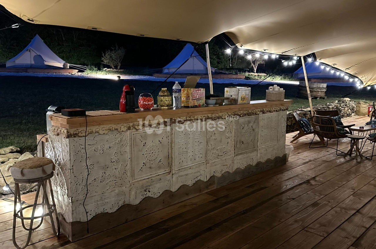 Comptoir en bois décoré sous une tente éclairée, avec des boissons et des collations, entouré de tables et de chaises.