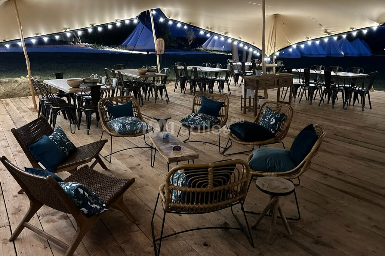 Espace extérieur couvert avec fauteuils en rotin, tables et chaises, éclairé par des guirlandes lumineuses la nuit.