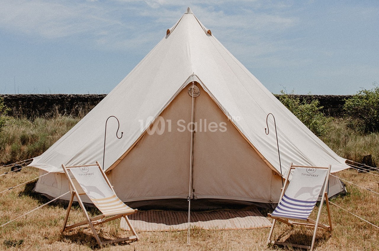 Tente en toile beige montée sur un terrain herbeux, entourée de deux chaises longues rayées.