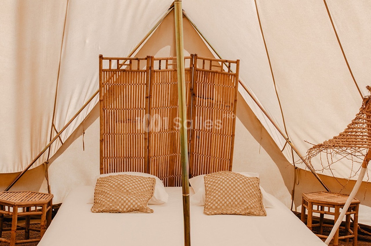 Lit double avec coussins beiges dans une tente équipée de cloisons en bambou et de petites tables en bois.