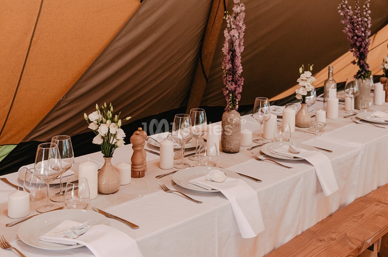 Table dressée avec nappes blanches, vaisselle élégante, bougies et fleurs dans un décor de tente.