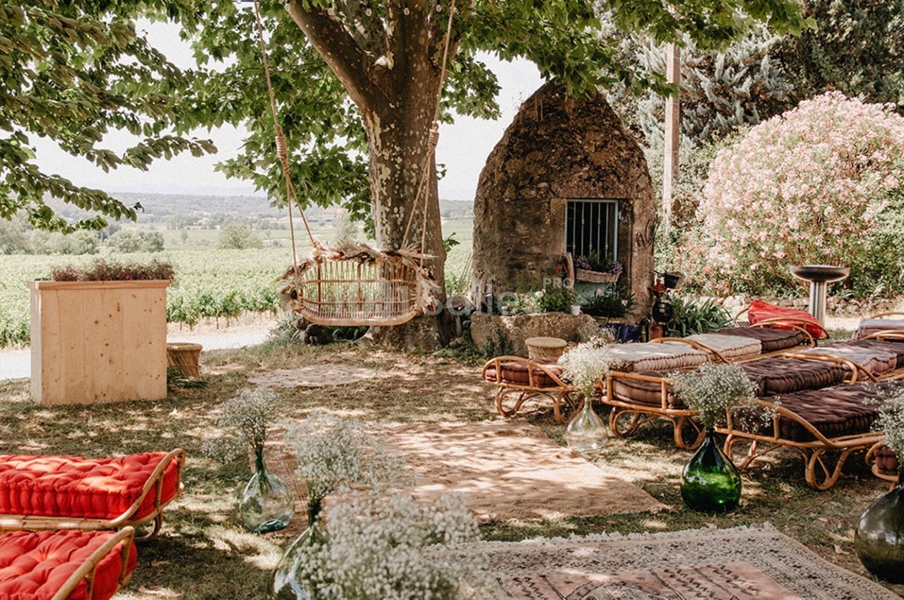 Espace extérieur aménagé avec des fauteuils en rotin, tapis, coussins et balançoire sous un arbre, près d'un paysage…