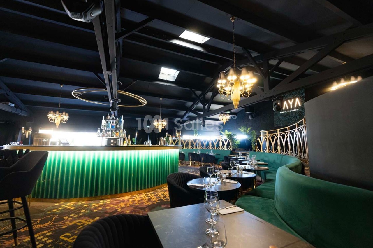 Intérieur d'un bar élégant avec comptoir lumineux vert, tables dressées et décorations modernes sous un plafond industriel.