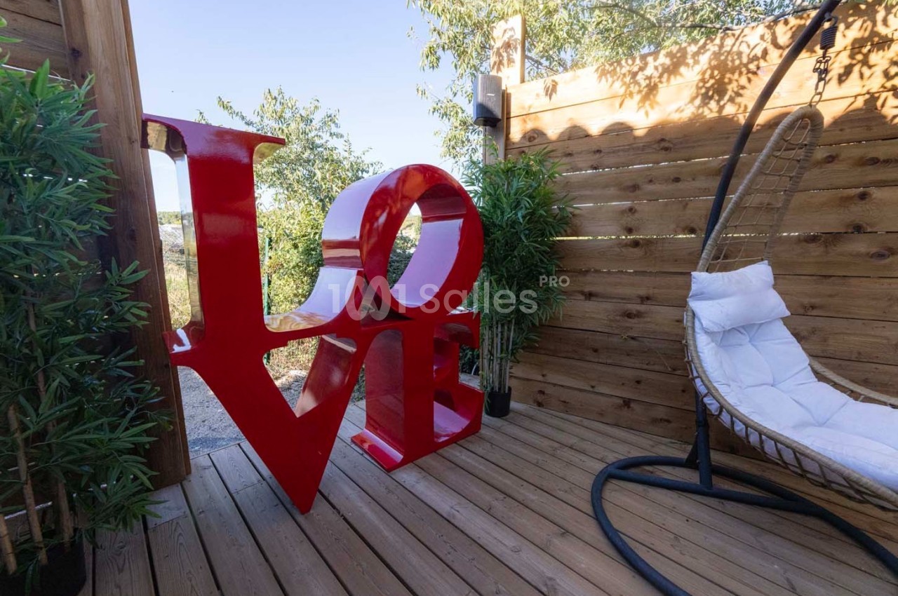 Sculpture rouge ’LOVE’ sur une terrasse en bois entourée de plantes et d'un fauteuil suspendu.