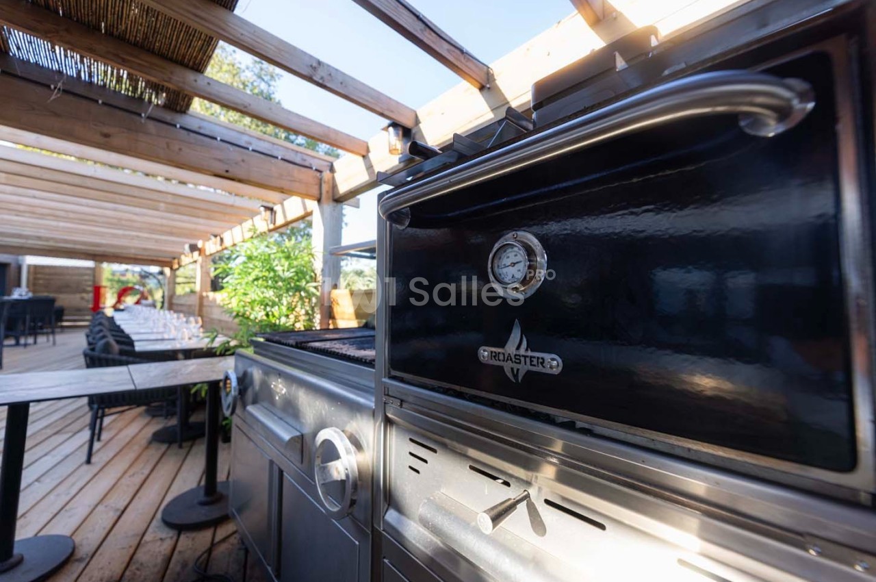Barbecue noir sur une terrasse en bois, entouré de tables et d'une pergola avec lumière naturelle.