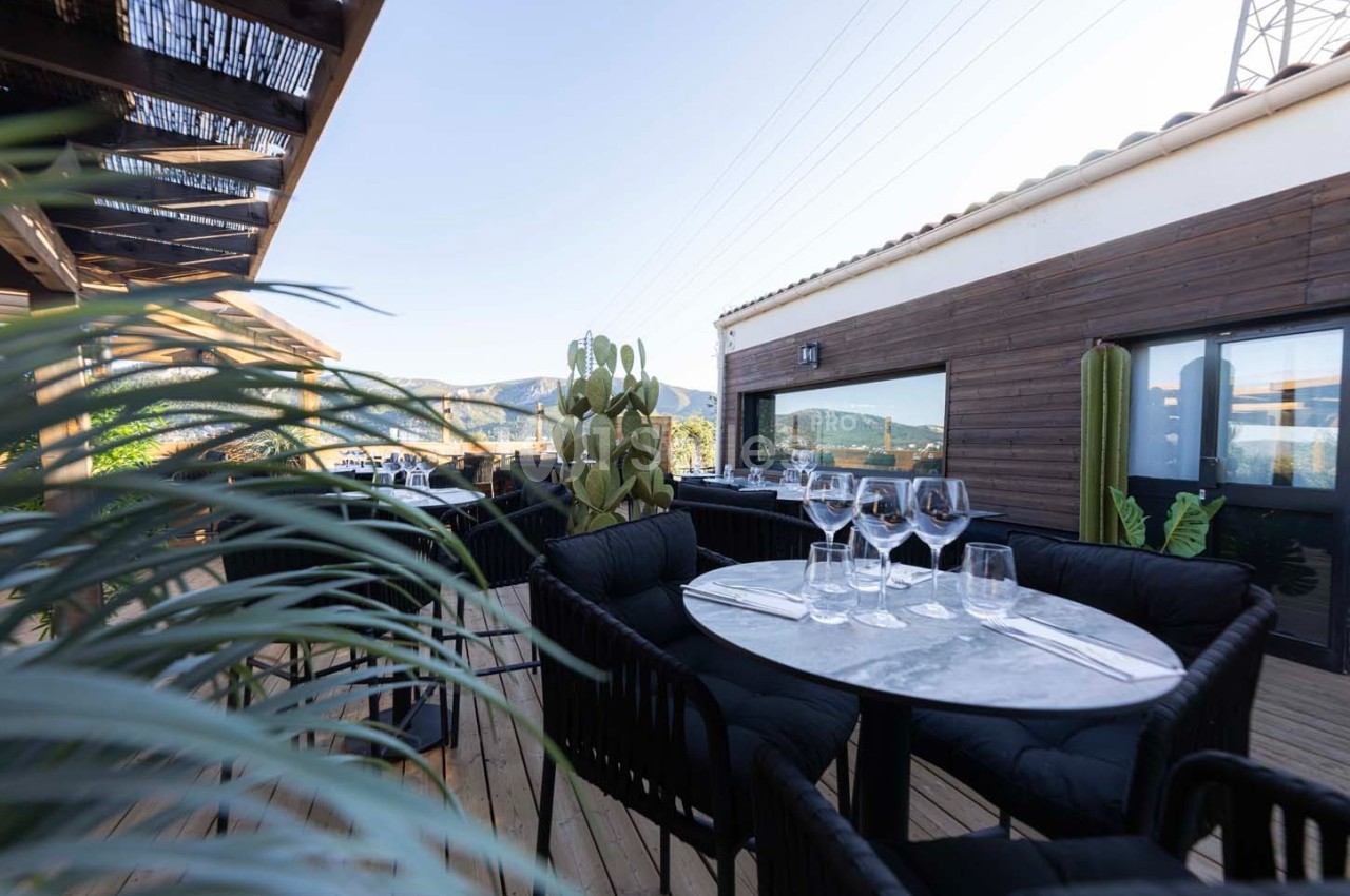 Terrasse en bois avec tables dressées, chaises noires, plantes et vue sur des collines sous un ciel dégagé.