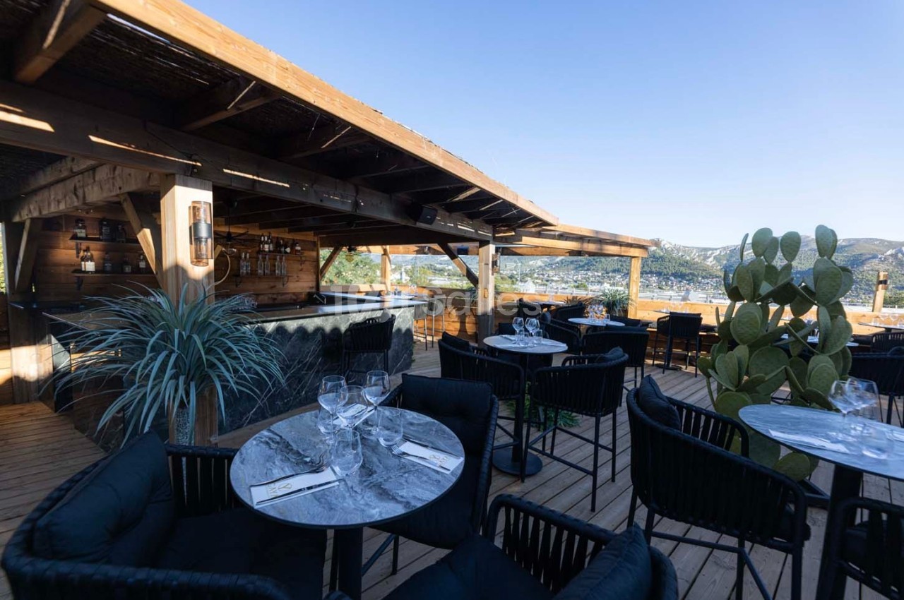 Terrasse en bois avec tables dressées, chaises noires, bar en arrière-plan et vue sur des collines ensoleillées.
