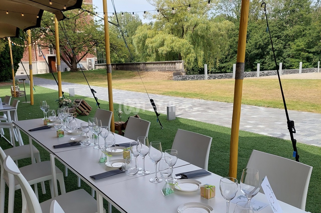 Table dressée en extérieur avec nappes blanches, verres et couverts, sous une tente avec vue sur un jardin verdoyant.