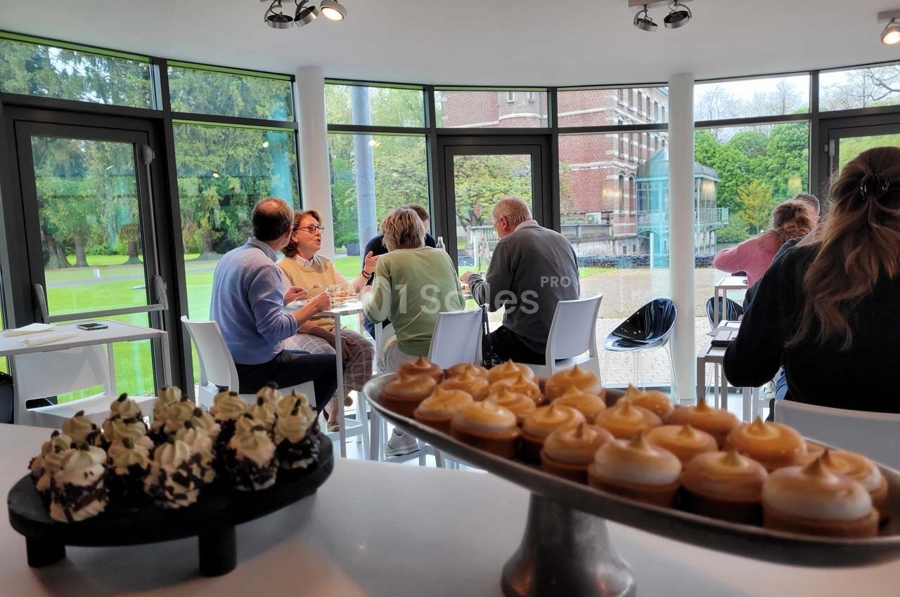 Des personnes discutent autour de tables dans une salle lumineuse avec vue sur un jardin, des desserts au premier plan.