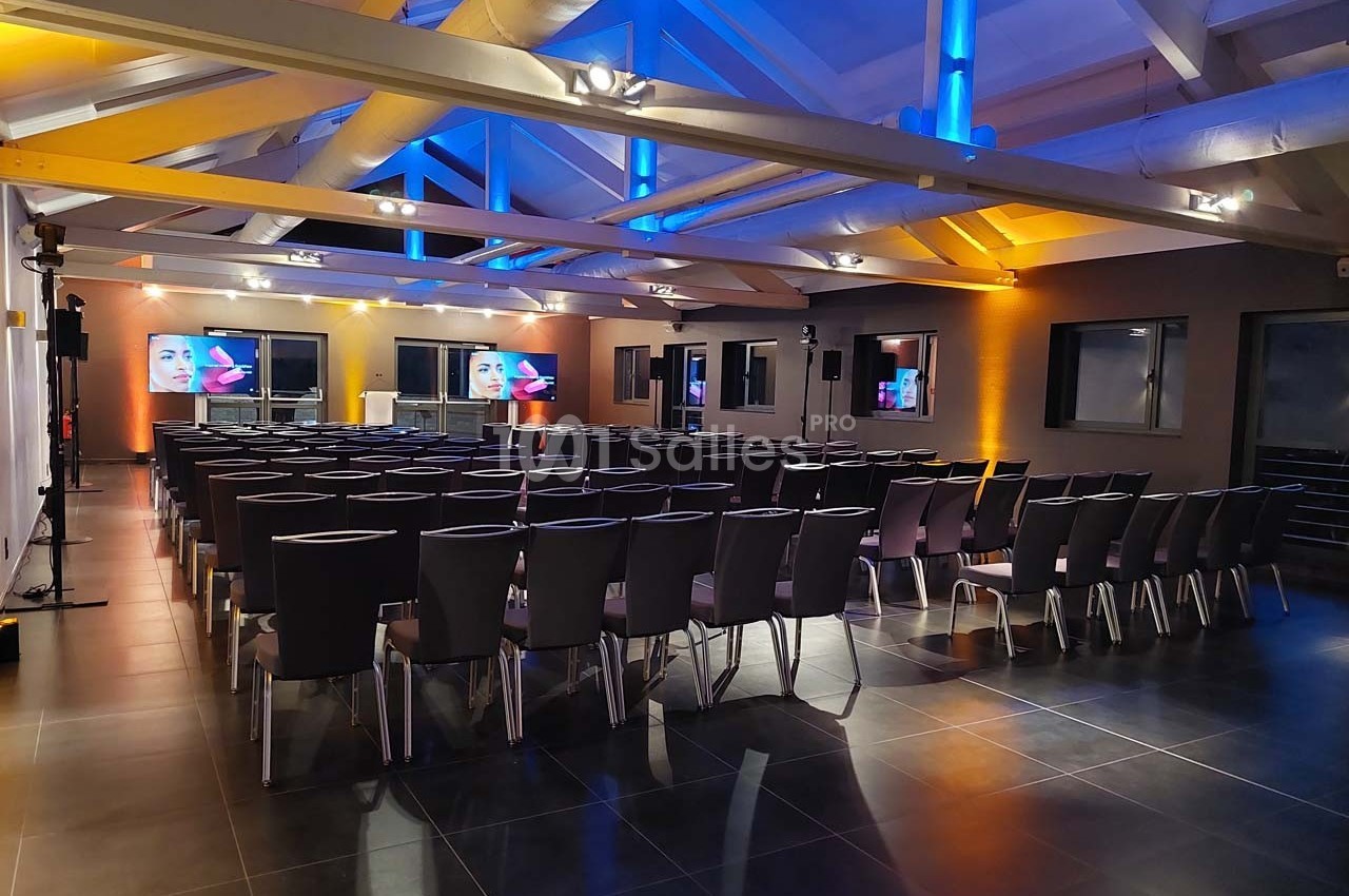 Salle de conférence moderne avec rangées de chaises noires, écrans allumés et éclairage coloré au plafond.