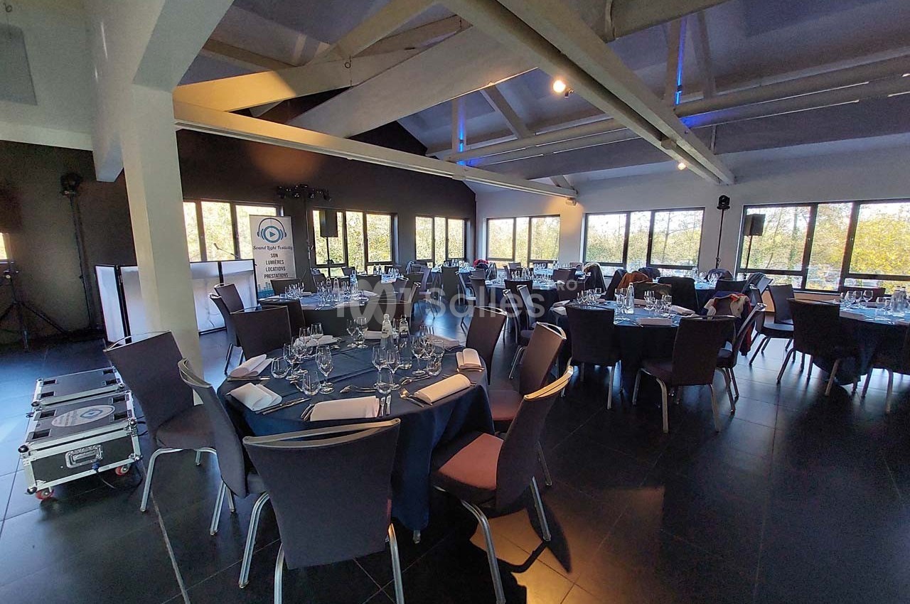 Salle de réception lumineuse avec tables rondes dressées, chaises modernes et grandes baies vitrées donnant sur l'extérieur.