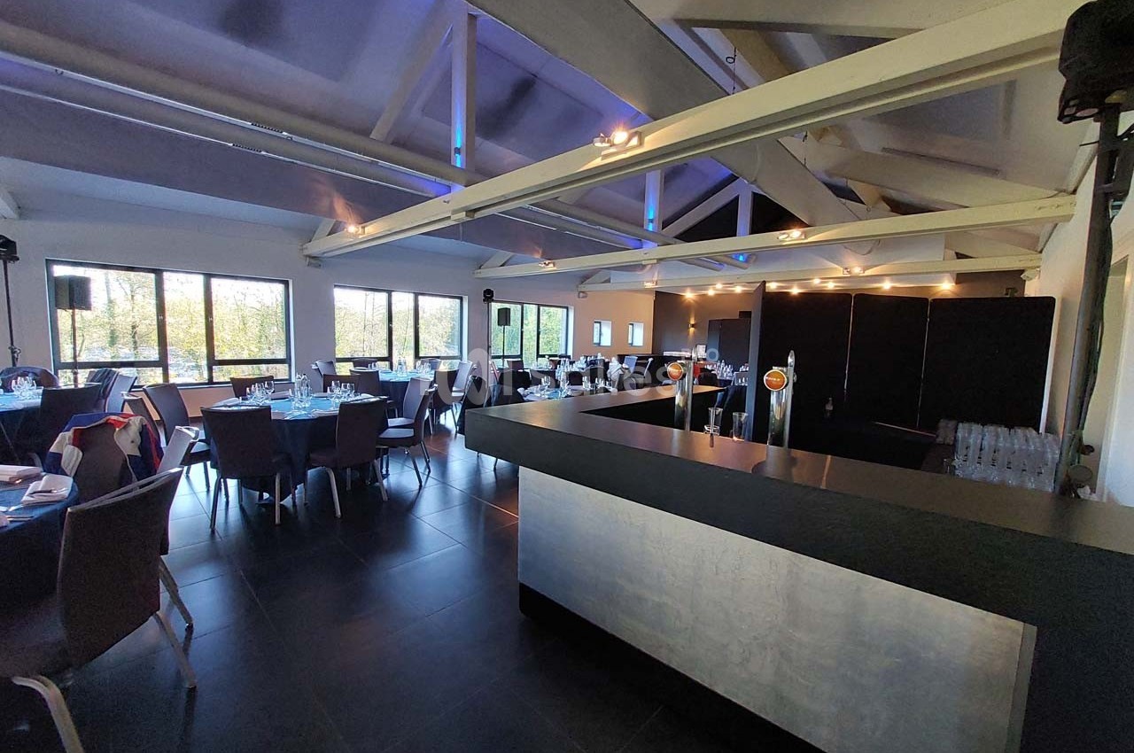 Salle moderne avec tables dressées, grandes baies vitrées, poutres apparentes et un comptoir au premier plan.