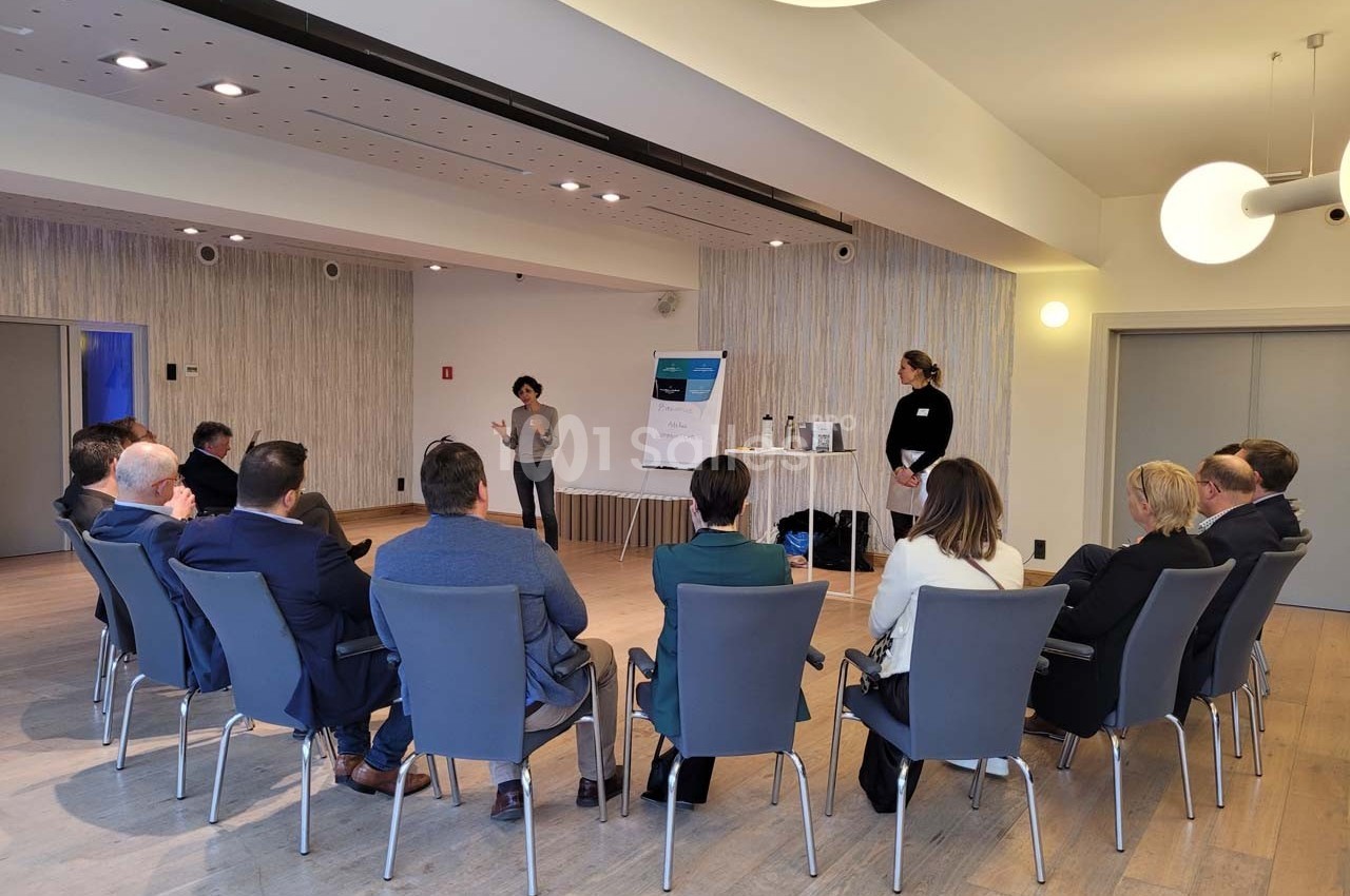 Une personne présente devant un groupe assis en cercle dans une salle lumineuse avec un tableau de conférence.