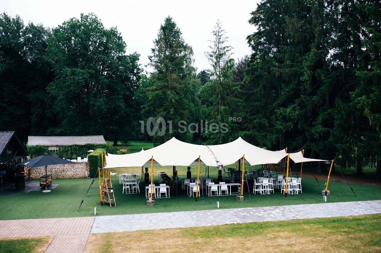 Tente blanche installée sur une pelouse entourée d'arbres, avec des tables et des chaises disposées en dessous.